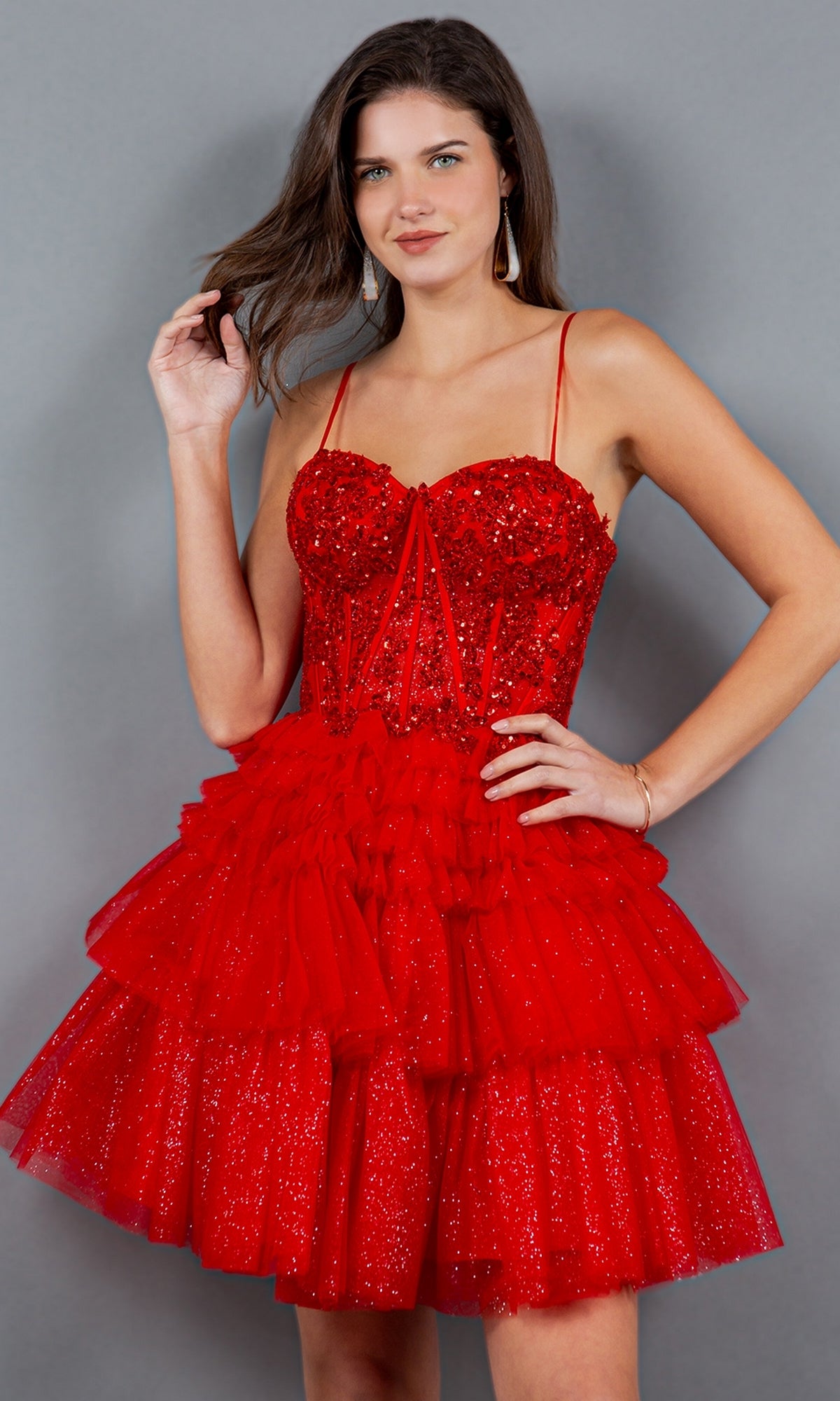 Woman wearing the Short Sweetheart Ruffled Homecoming Dress 5133J, featuring sparkly red fabric, thin straps, and a short ruffled skirt, standing against a grey background.